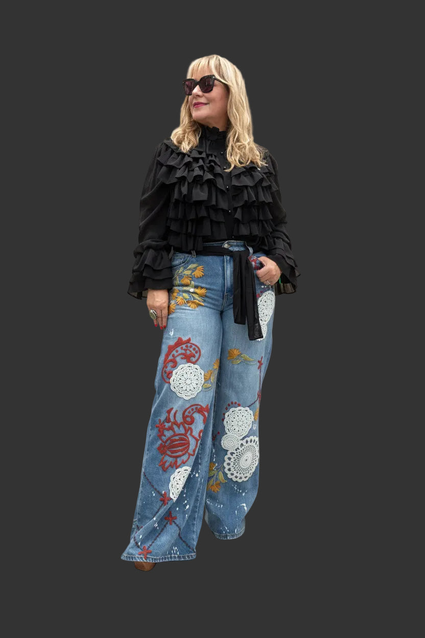 A woman modeling a black ruffled long-sleeve top and wide-leg blue denim jeans decorated with colorful red, yellow, and white floral patterns, standing against a beige studio backdrop.
