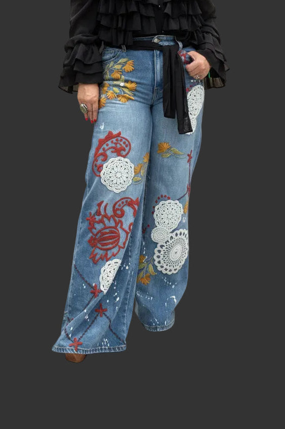 A woman modeling a black ruffled long-sleeve top and wide-leg blue denim jeans decorated with colorful red, yellow, and white floral patterns, standing against a beige studio backdrop.