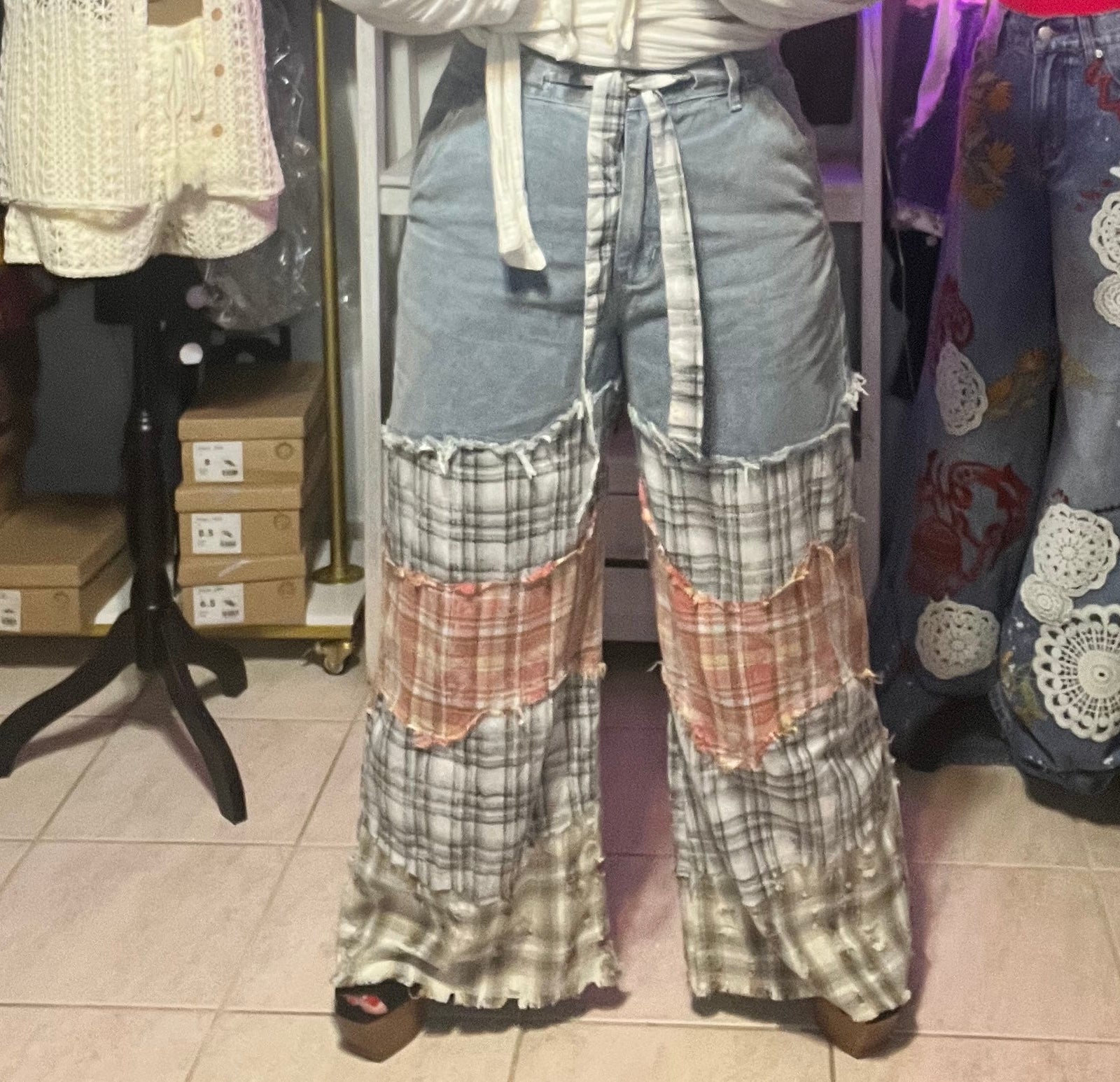A woman modeling a white cropped hoodie with a drawstring, paired with light-wash jeans featuring crochet details and plaid cropped pants, standing in a clothing store.